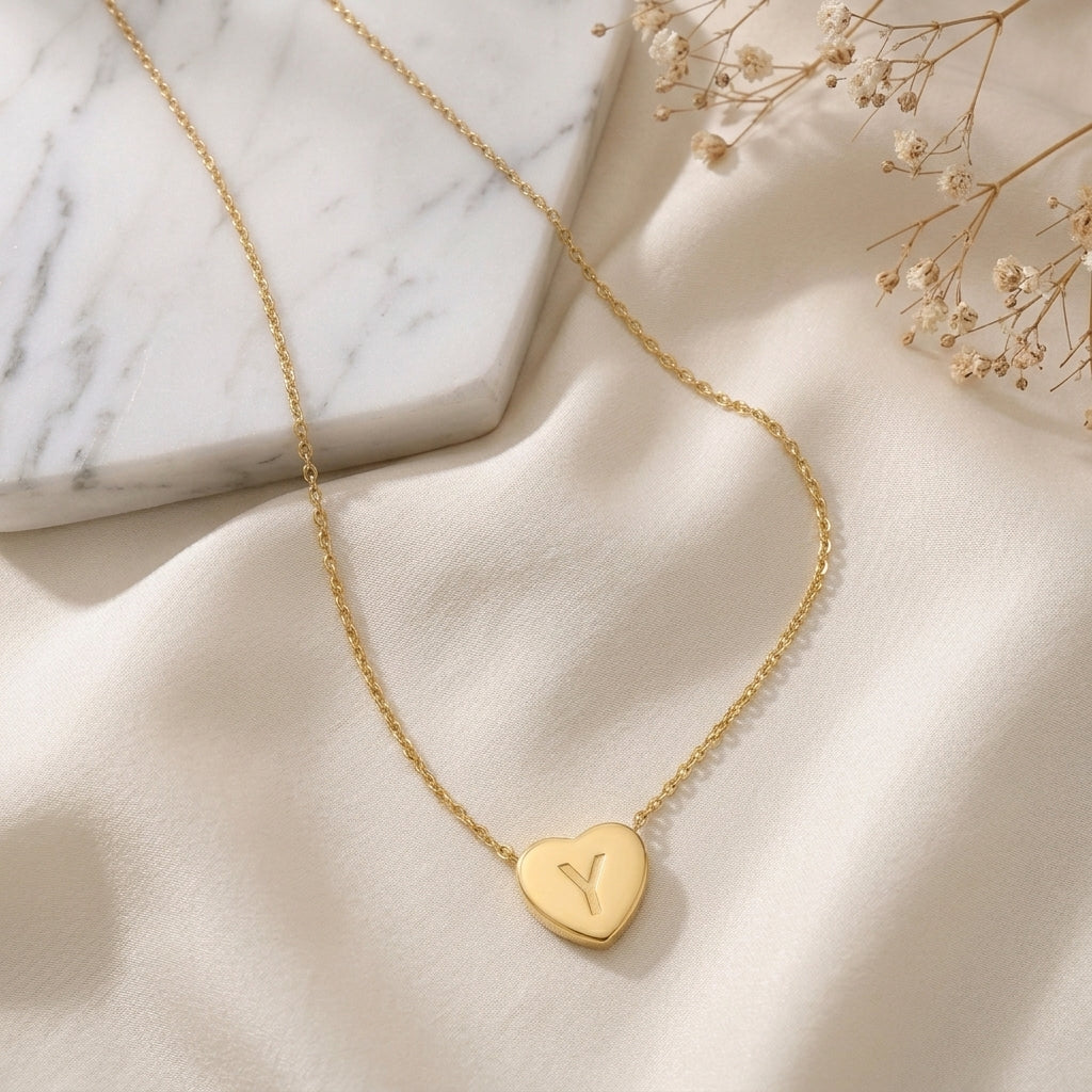 Collier doré à petit cœur gravé d'une initiale, posé à plat sur un tissu clair