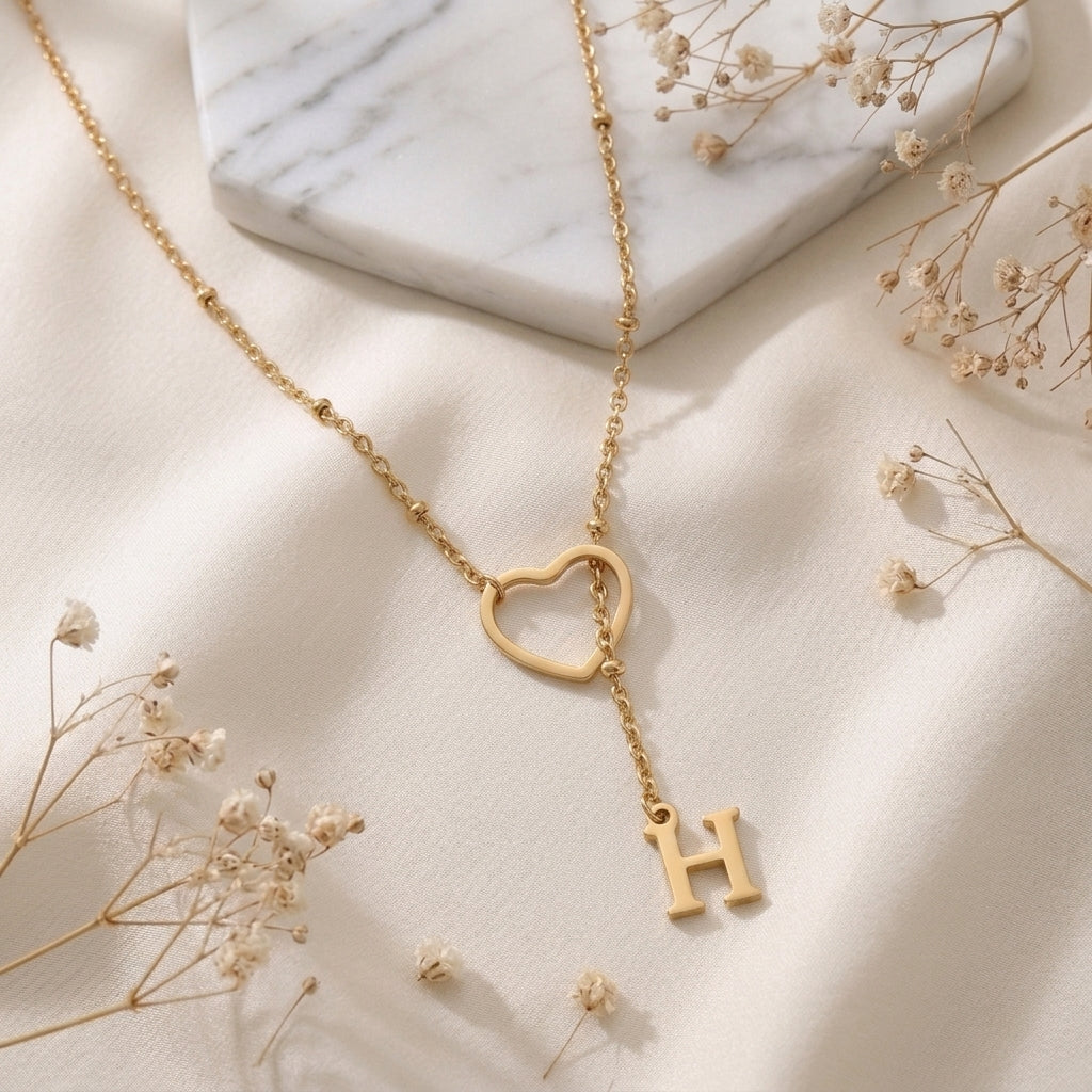 Collier doré à cœur ajouré et initiale posé à plat sur un tissu clair