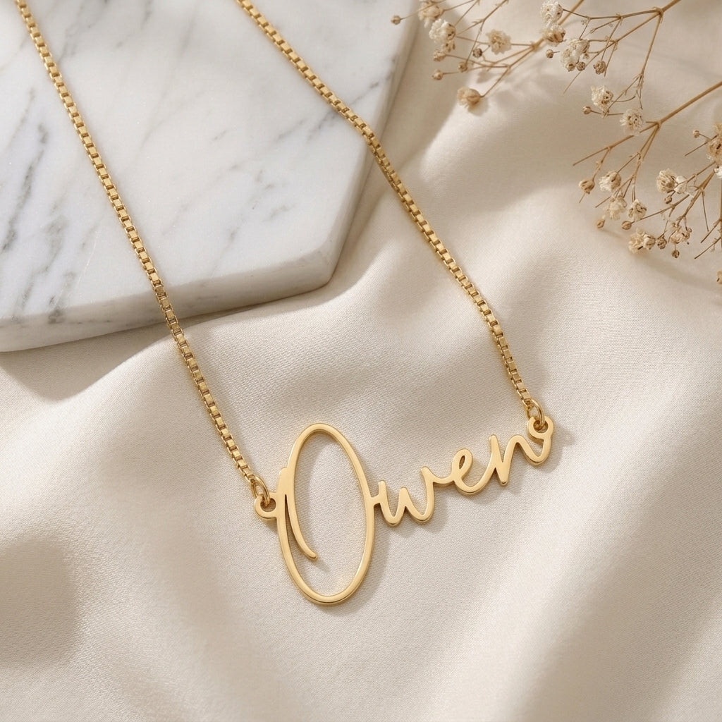 Collier prénom doré en écriture manuscrite posé à plat sur un tissu clair