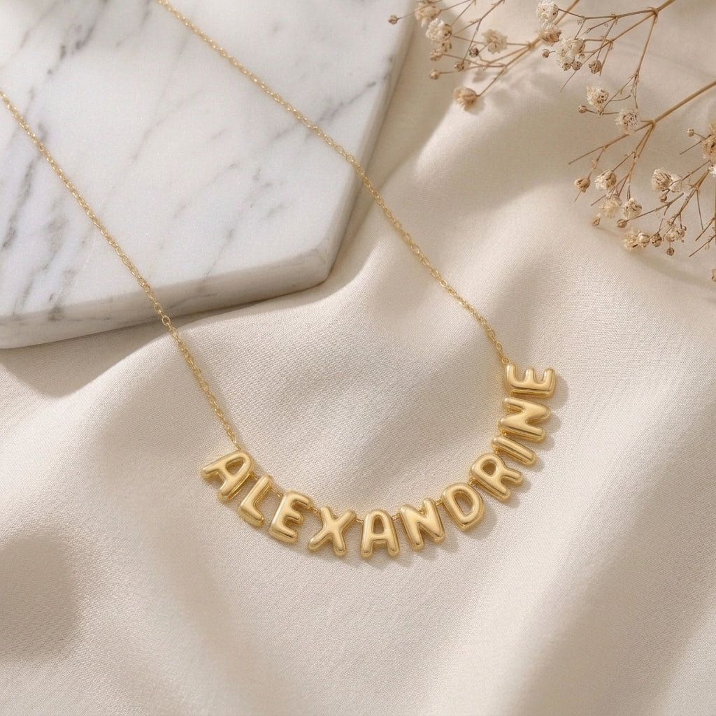 Collier doré à lettres bombées formant un prénom, posé à plat